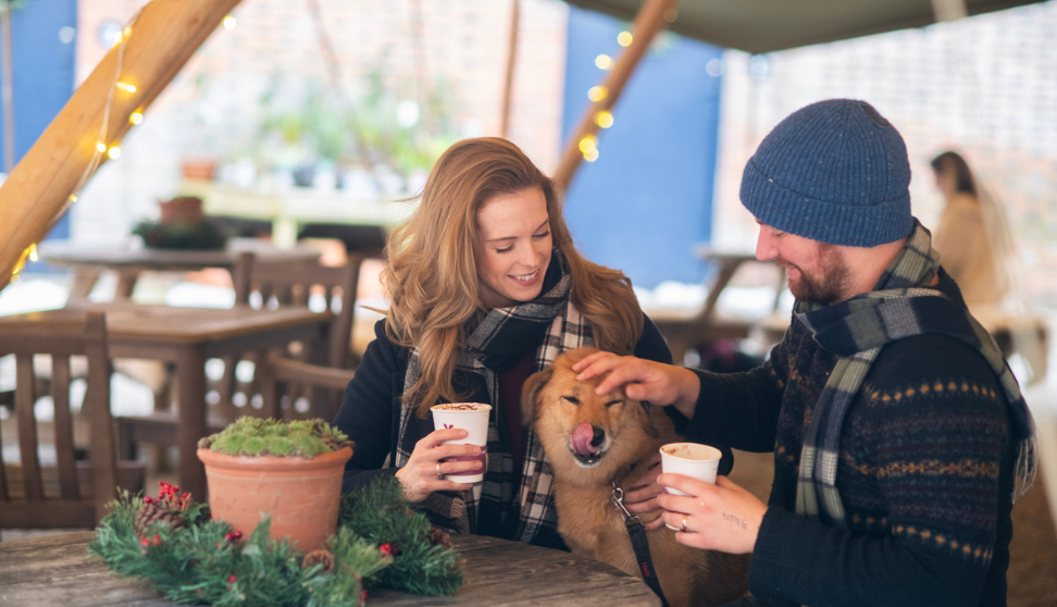 Two people enjoying hot drinks at an outdoor café with their dog, wearing winter scarves and surrounded by festive decorations.